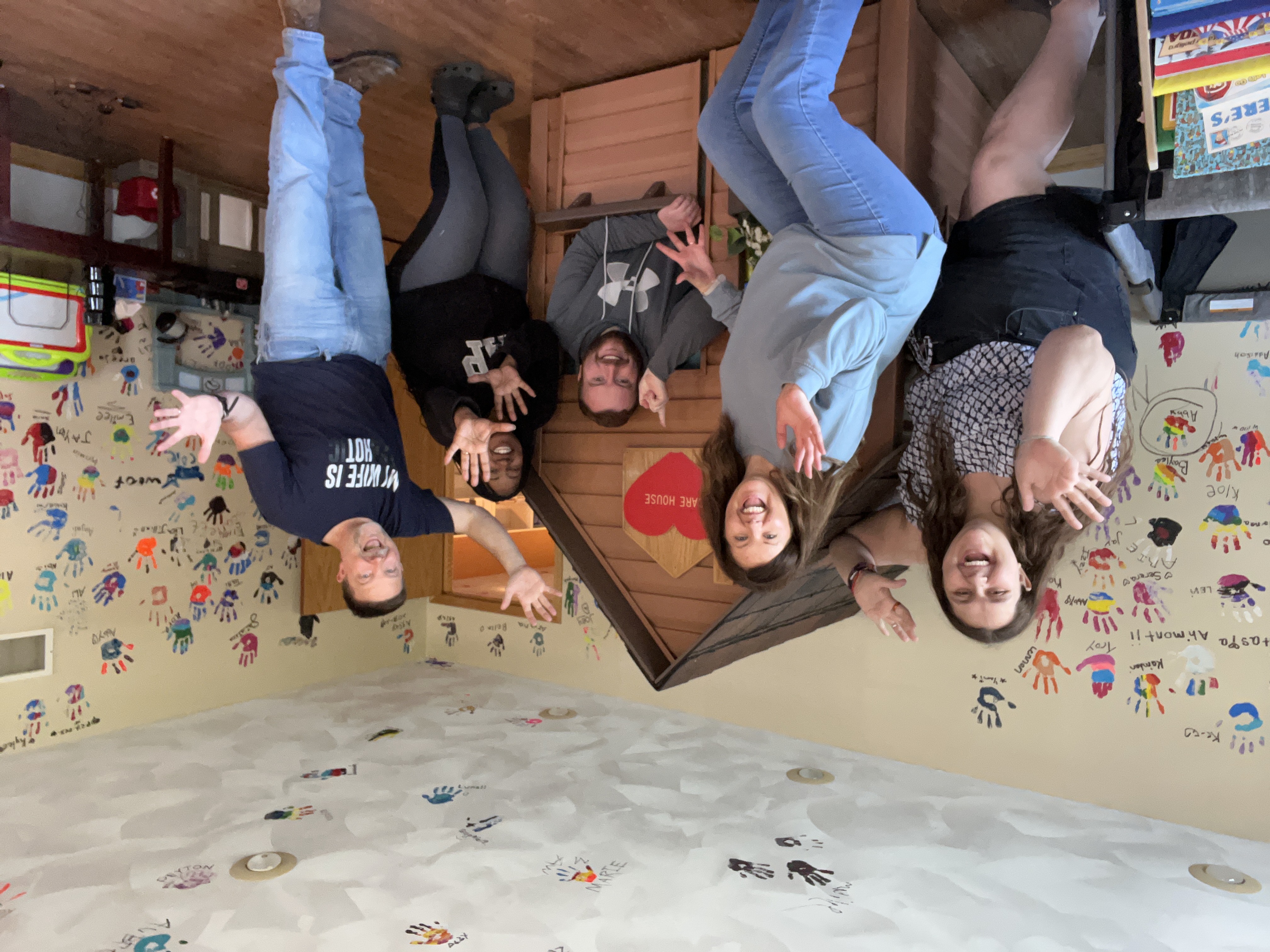 A group of 5 people look excited with jazz hands standing around a small play house with a heart shaped sign saying 